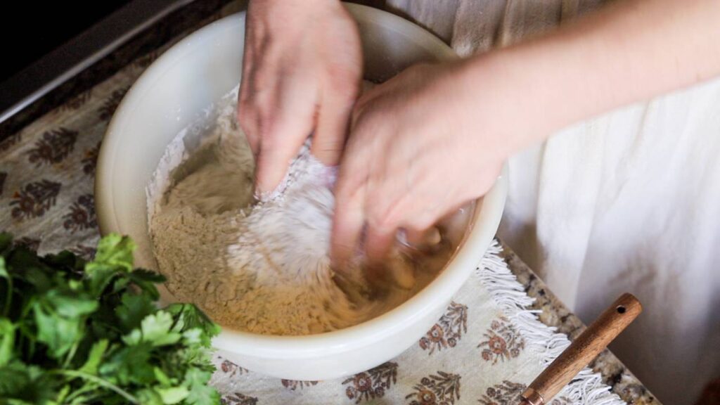 Hands mixing flour and water in a white bowl to start sourdough dough.
