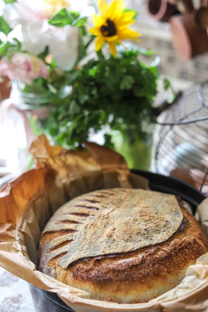 Sourdough bread in a Dutch oven lined with parchment paper, sitting on a counter with flowers in the background.