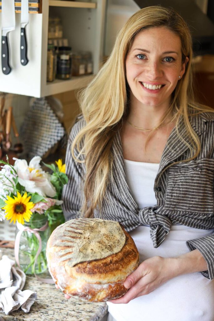 Woman smiling and holding a round loaf of sourdough bread in a cozy kitchen.