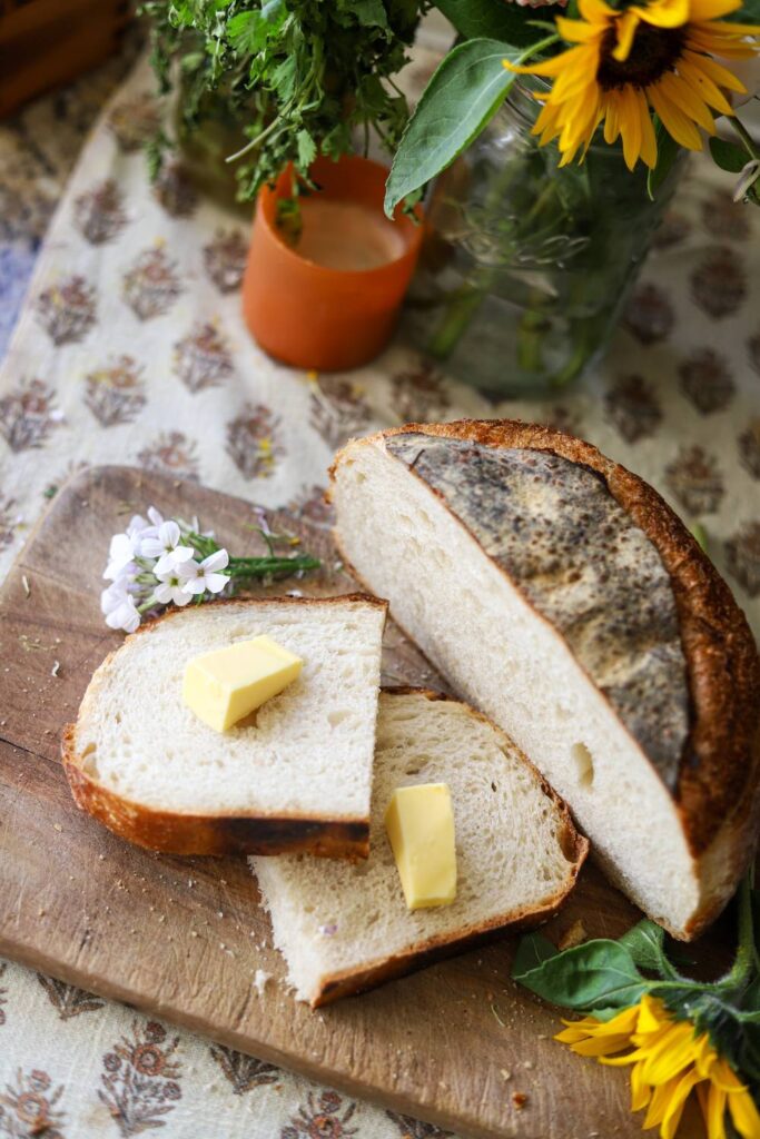 Top-down view of sliced sourdough bread with butter, surrounded by sunflowers and fresh herbs.