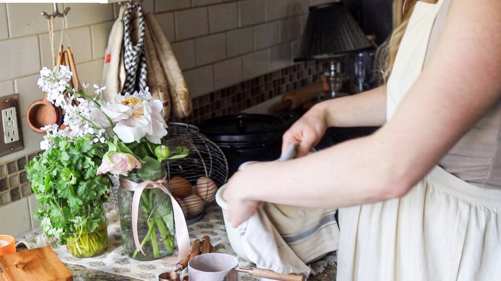 Woman covering a loaf of dough with a cloth next to a vase of flowers and fresh herbs.