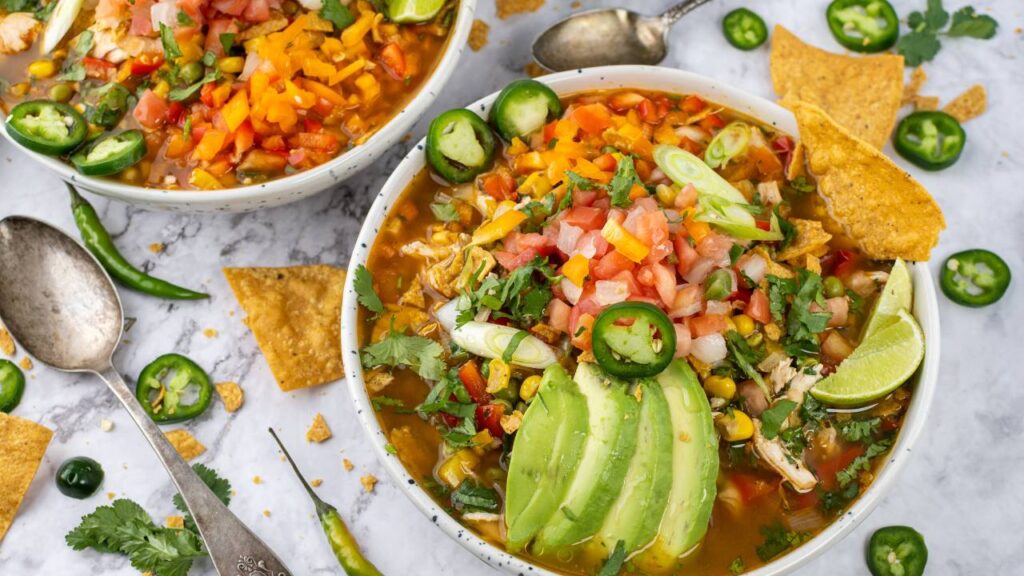 Close-up of chicken tortilla soup with avocado slices, diced vegetables, cilantro, jalapeños, and lime.