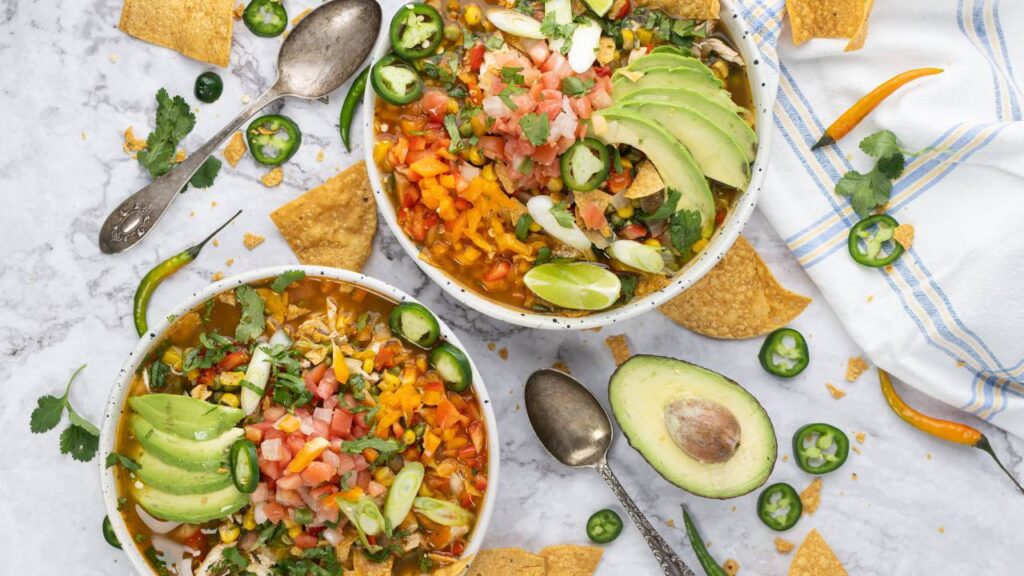 Two bowls of chicken tortilla soup on a marble surface with avocado, peppers, lime, and tortilla chips around them.