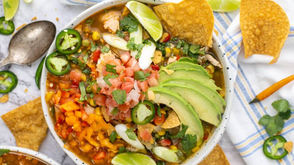 Overhead view of loaded chicken tortilla soup with avocado, pico de gallo, lime wedges, jalapeños, and tortilla chips.