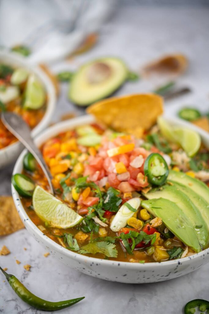 Bowl of chicken tortilla soup topped with avocado slices, diced tomatoes, jalapeños, lime, and tortilla chips.
