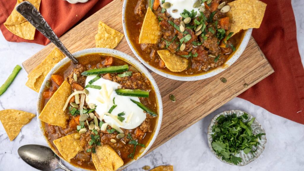Two bowls of pumpkin chili topped with sour cream, pumpkin seeds, herbs, and tortilla chips on a wooden board.