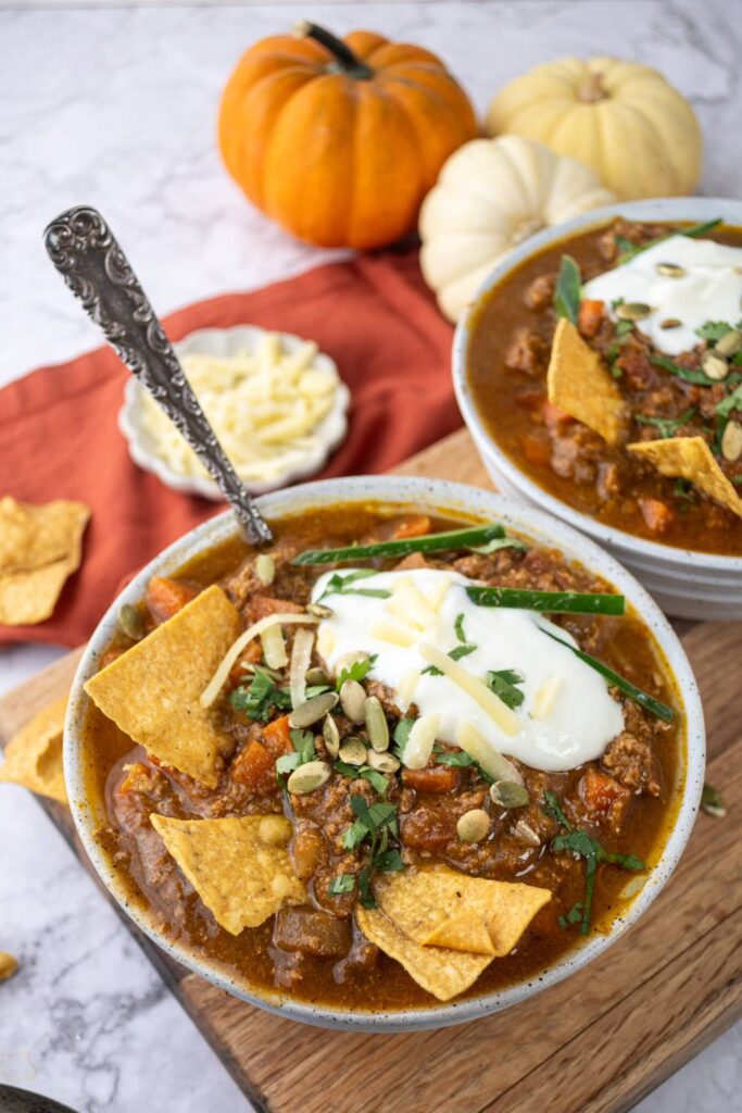 Bowl of pumpkin chili topped with sour cream, cheese, herbs, and tortilla chips with pumpkins in the background.