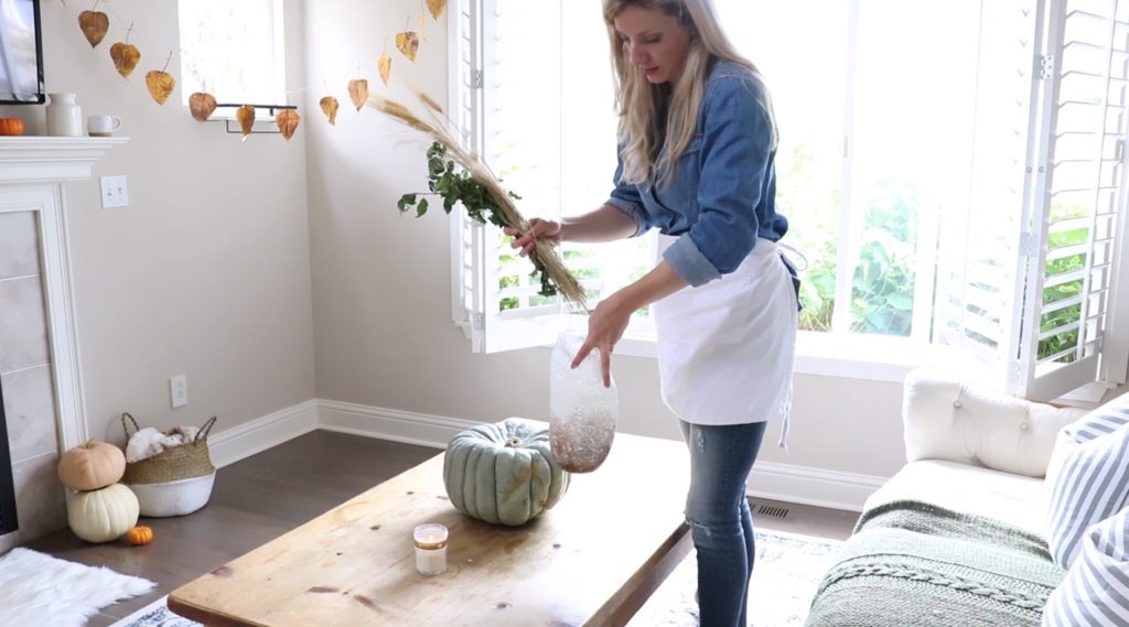 girl putting fall florals in a vase with hanging leaves in the background