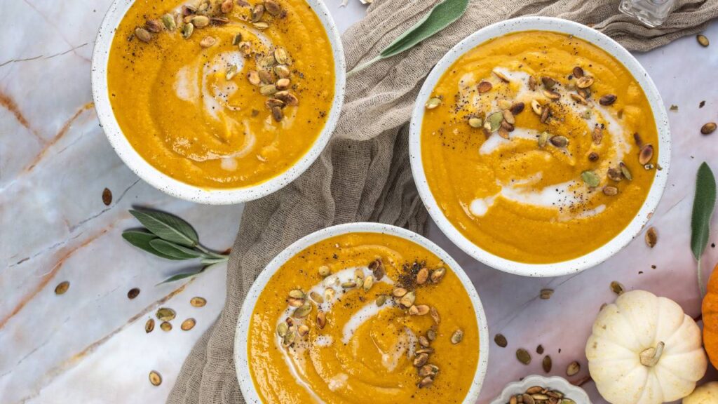 Three bowls of butternut squash soup on a table surrounded by sage leaves and small pumpkins.