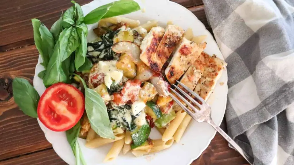 creamy basil chicken pasta on a white plate with a garnish of roma tomato and fresh basil on a farmhouse table with gray and white checked cloth