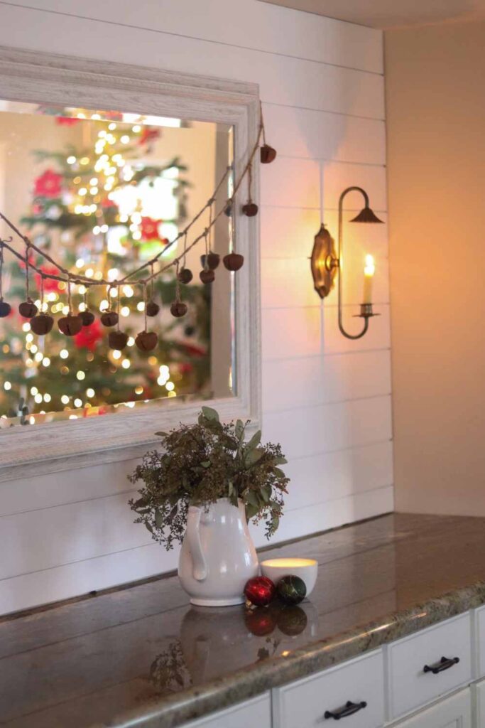 Buffet decorated for Christmas with sleigh bells, a white pitcher of greenery, ornaments, and brass sconce on a shiplap wall