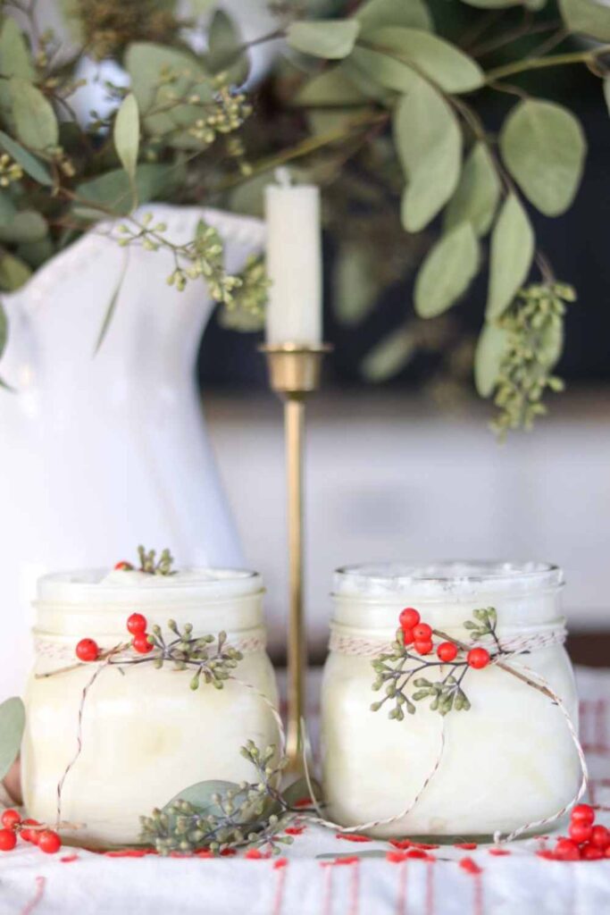 Two jars of DIY homemade Christmas body butter decorated with bakers twine and sprigs of juniper berry, with a brass candlestick in the background