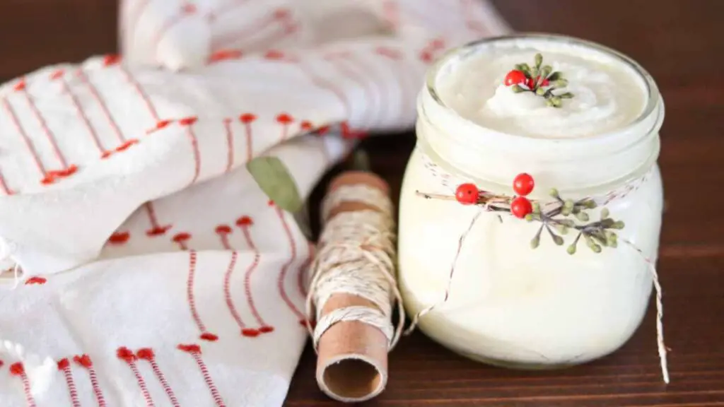 a jar of homemade Christmas juniper berry body butter, a roll of bakers twine, and a red and white cloth, on a farmhouse table