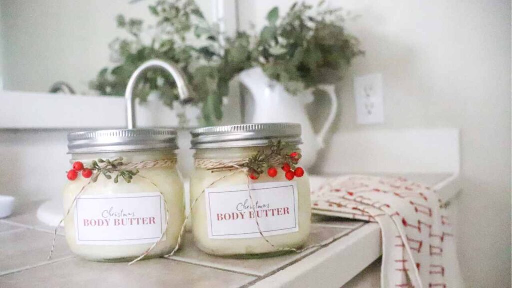 Two jars of homemade Christmas body butter with printed labels, decorated with bakers twine and sprigs of juniper, on a bathroom countertop