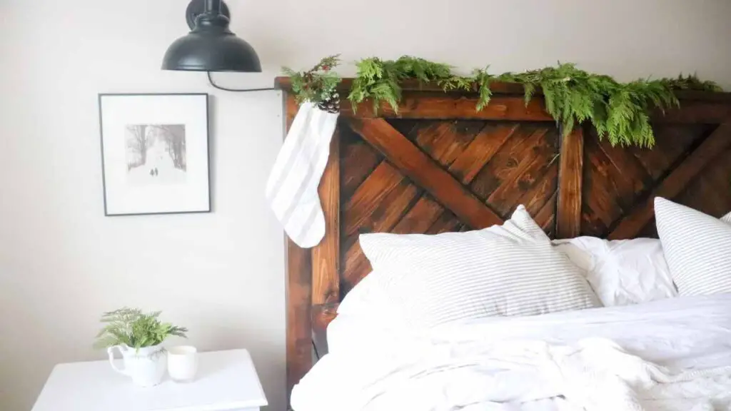 rustic farmhouse bed with fluffy white bedding, headboard decorated for Christmas with pine garland and white stockings with pinecones