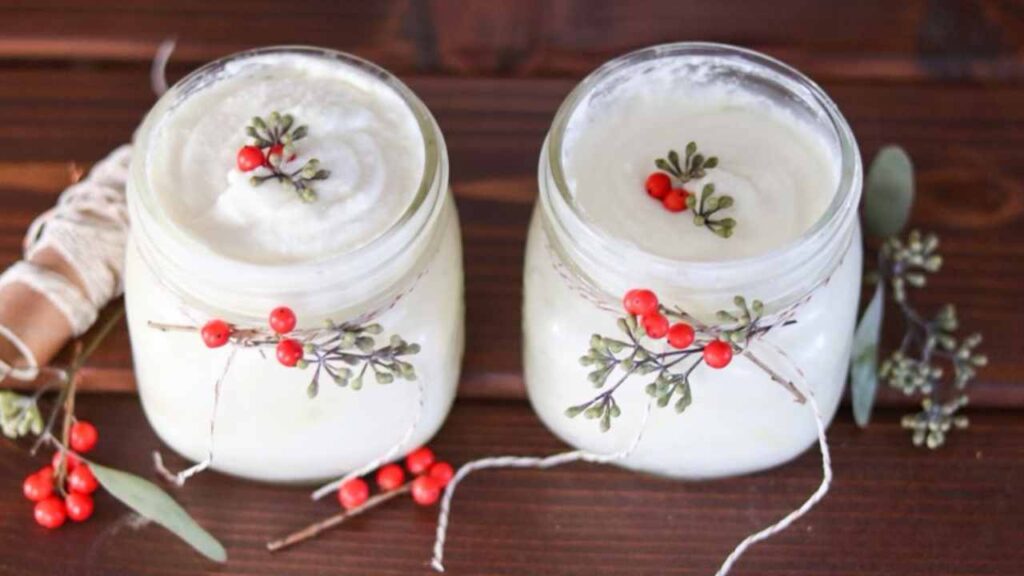 Two jars of homemade Christmas juniper berry body butter, decorated with bakers twine and juniper sprigs, on a farmhouse table