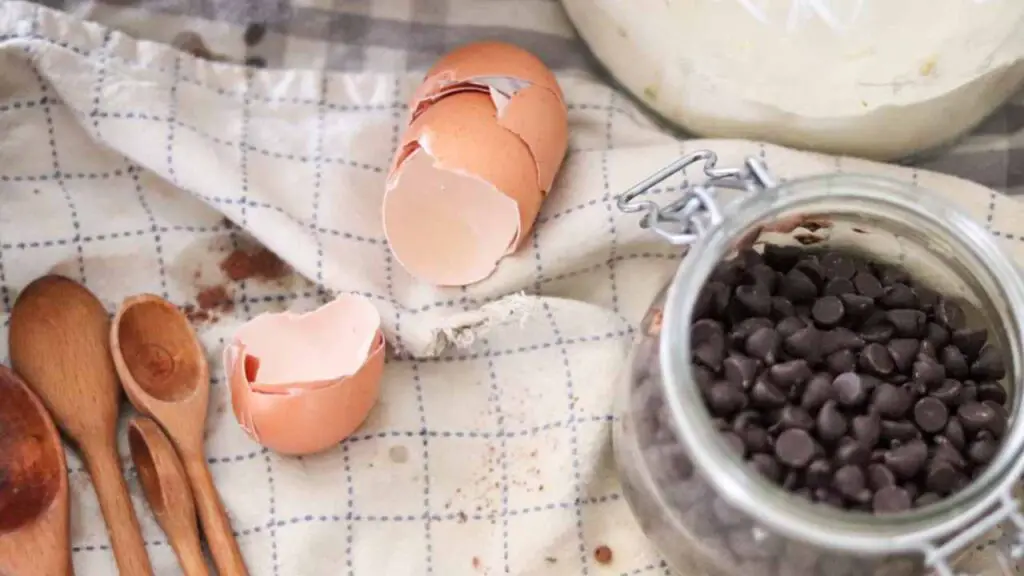 cracked brown eggshells and a jar of chocolate chips, with wooden baking utensils, atop a blue and white cloth