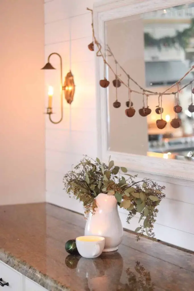 Buffet decorated for Christmas with sleigh bells, a white pitcher of greenery, and brass sconce on a shiplap wall