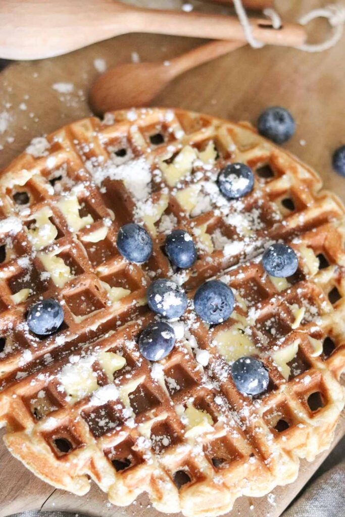 Homemade einkorn waffle with butter, powdered sugar, and fresh blueberries on a wood serving board with wooden spoons