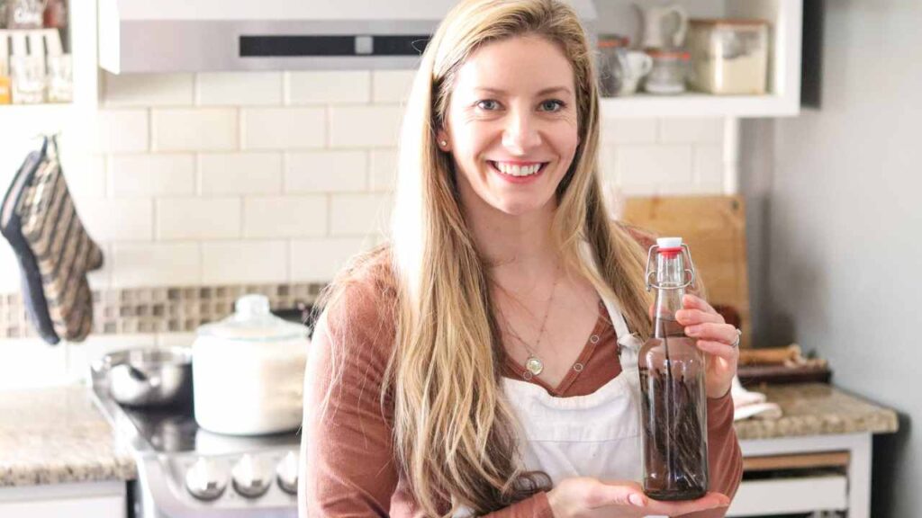 Woman standing in a farmhouse kitchen holding a bottle of homemade vanilla extract