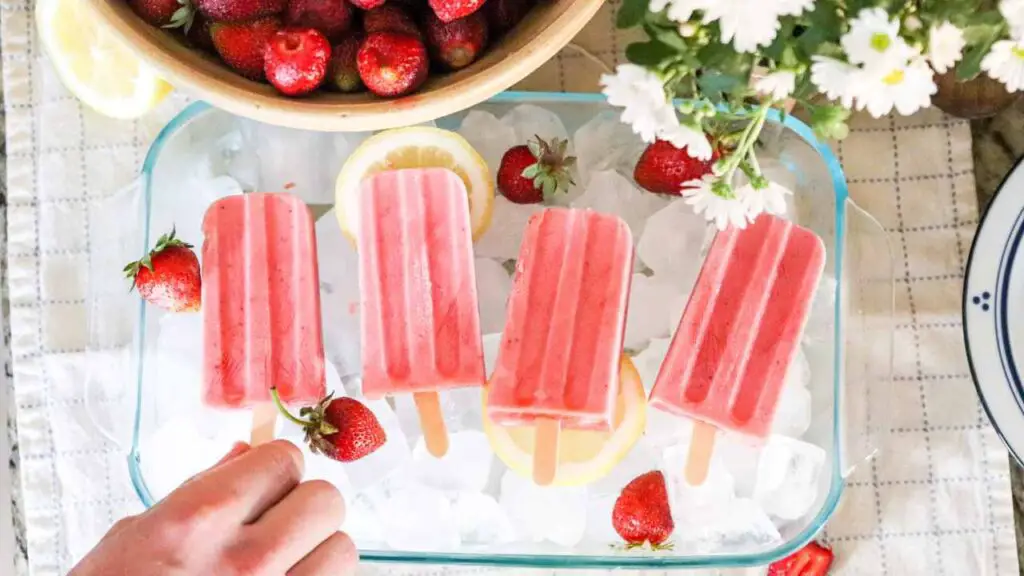 hand grabbing a strawberry cream kefir popsicle in a dish of ice with strawberries and daisies