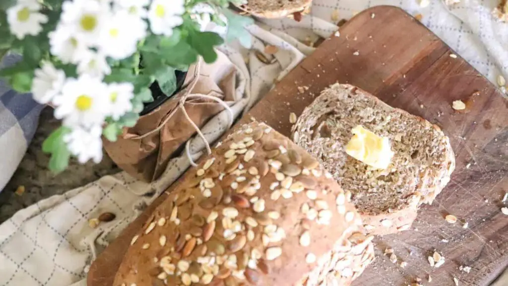 a loaf of homemade whole wheat seeded sourdough sandwich bread on a wood cutting board with farmhouse cloth and daisies
