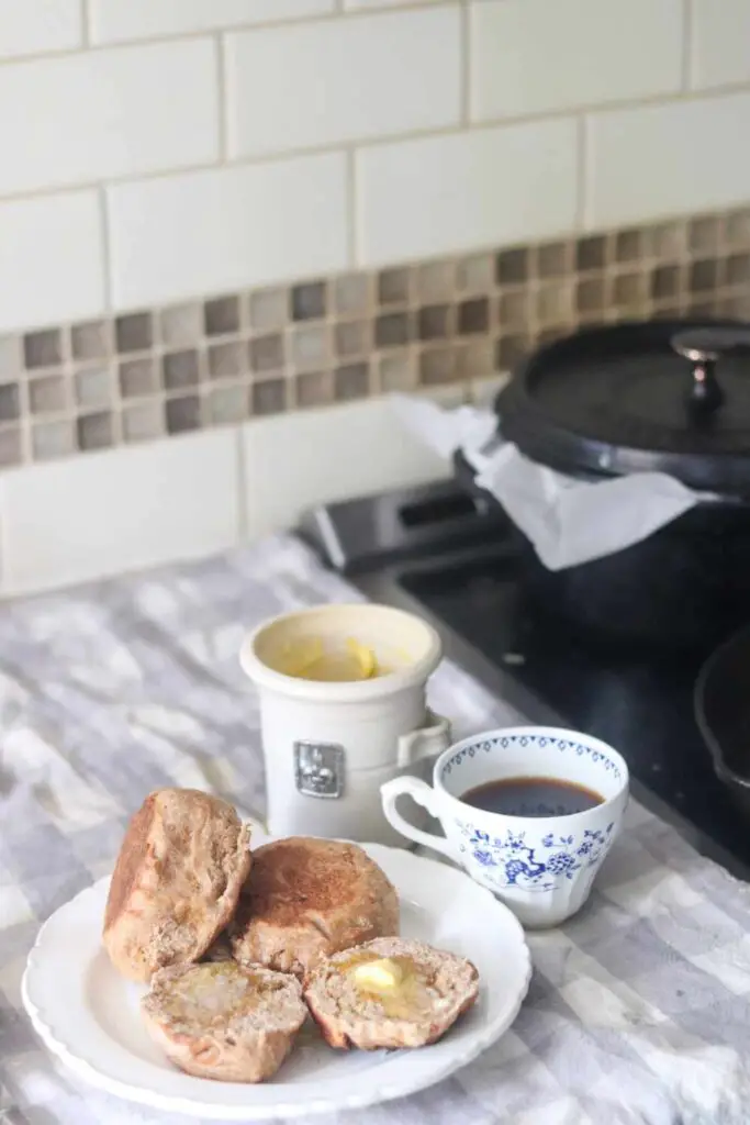 whole wheat sourdough English muffins topped with honey butter on a white plate, next to a blue and white china cup of coffee, on a checked towel