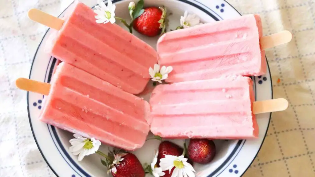 strawberry cream kefir popsicles in a white and blue bowl with daisies and strawberries on a checked cloth