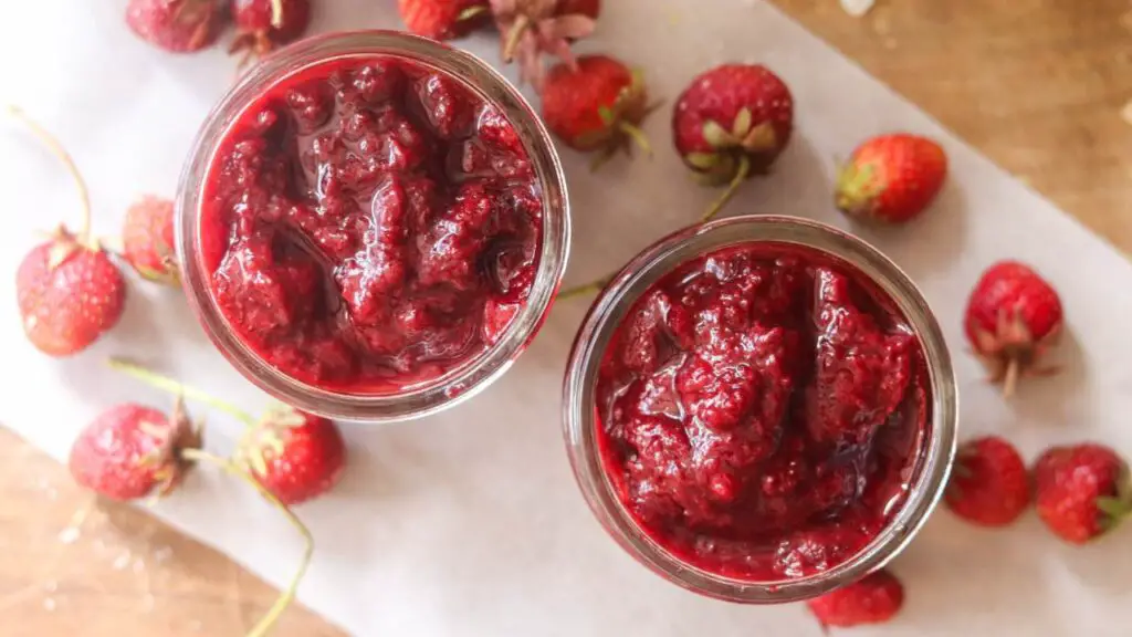 Two jars of strawberry jam with fresh strawberries around them.