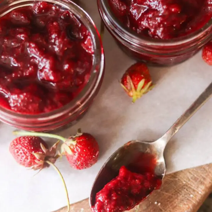 Two jars of strawberry jam with a spoonful of strawberry jam and fresh strawberries.