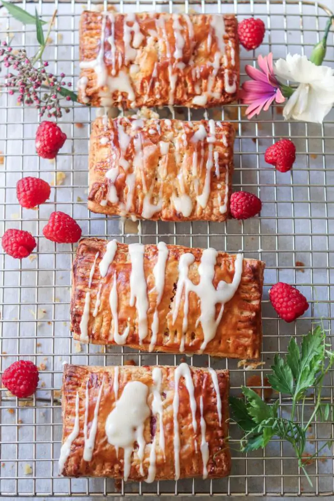 A cooling rack with homemade raspberry pop tarts on it with fresh raspberries and flowers. 