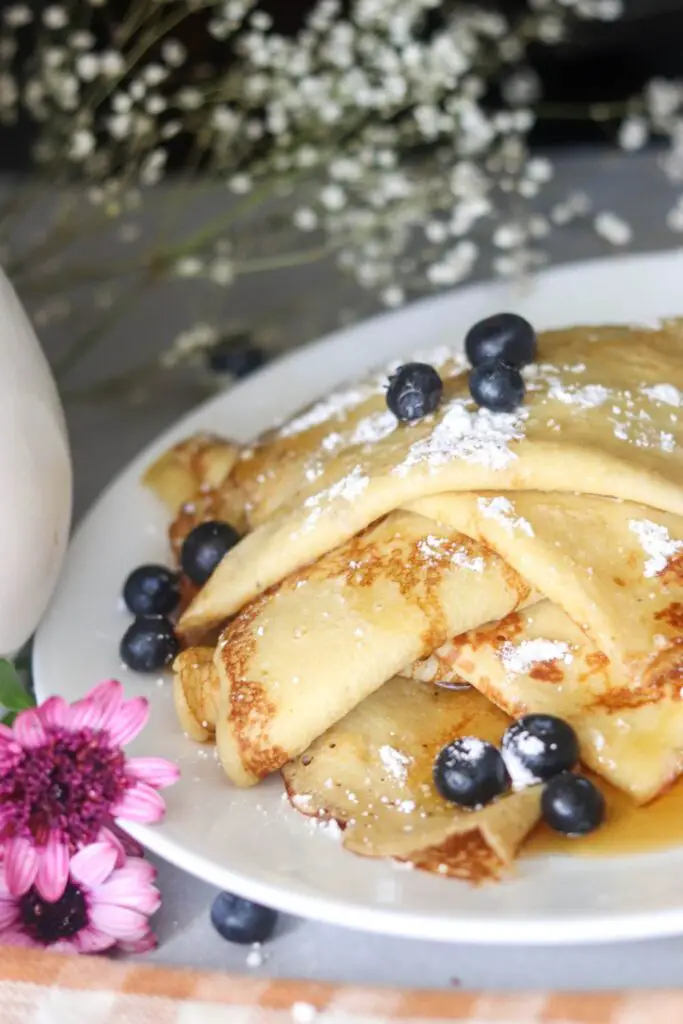 A white plate of sourdough crepes with fresh blueberries and pink flowers.