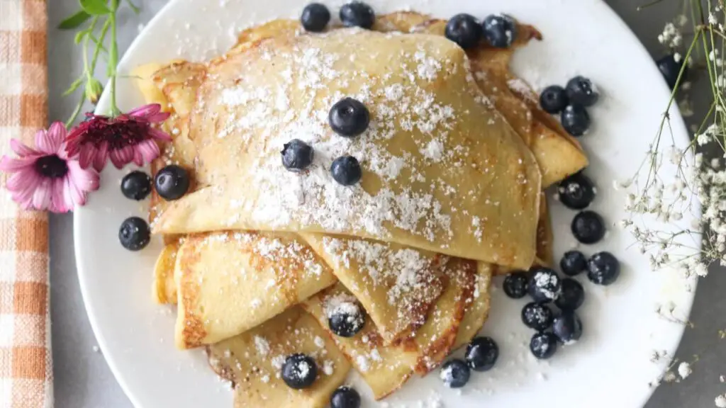 A plate of sourdough crepes with fresh blueberries and powdered sugar on top.