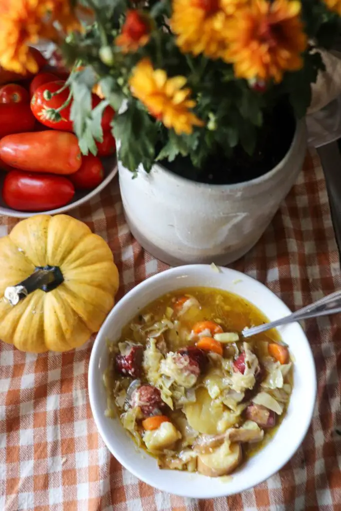 A bowl of bratwurst soup with a pumpkin, bowl of tomatoes, and fresh flowers.