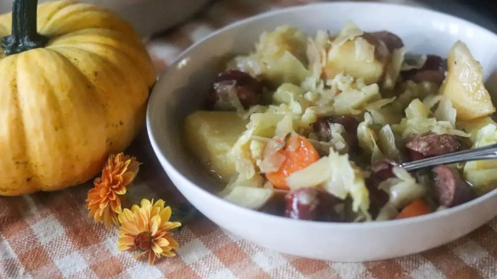 A bowl of bratwurst soup next to a pumpkin and orange flowers.