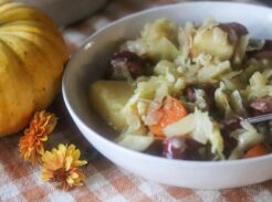 A bowl of bratwurst soup next to a pumpkin and orange flowers.