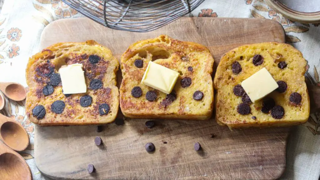 Three pieces of french toast on a wooden cutting board with butter and chocolate chips.