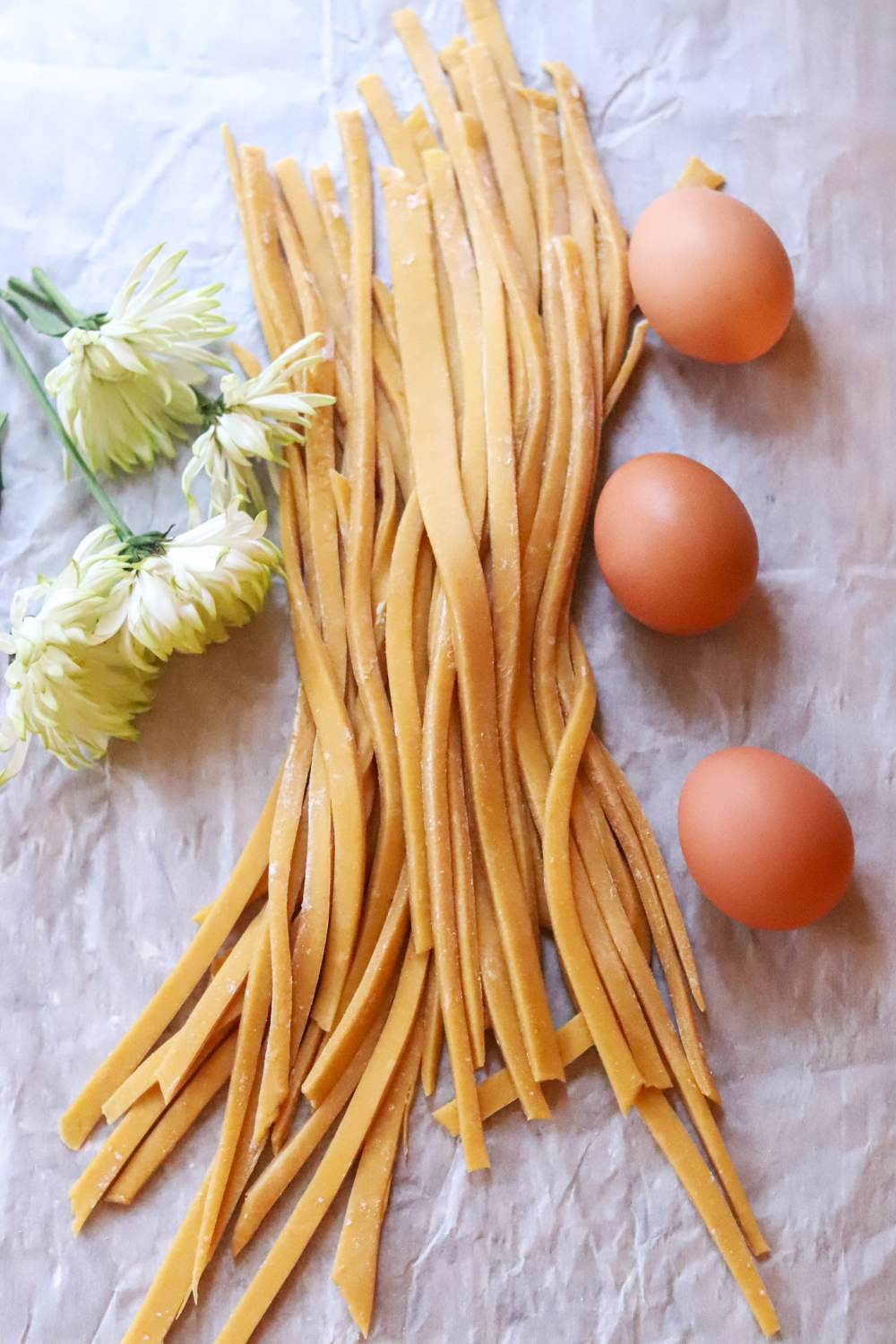 Homemade einkorn pasta on a table with eggs and flowers.