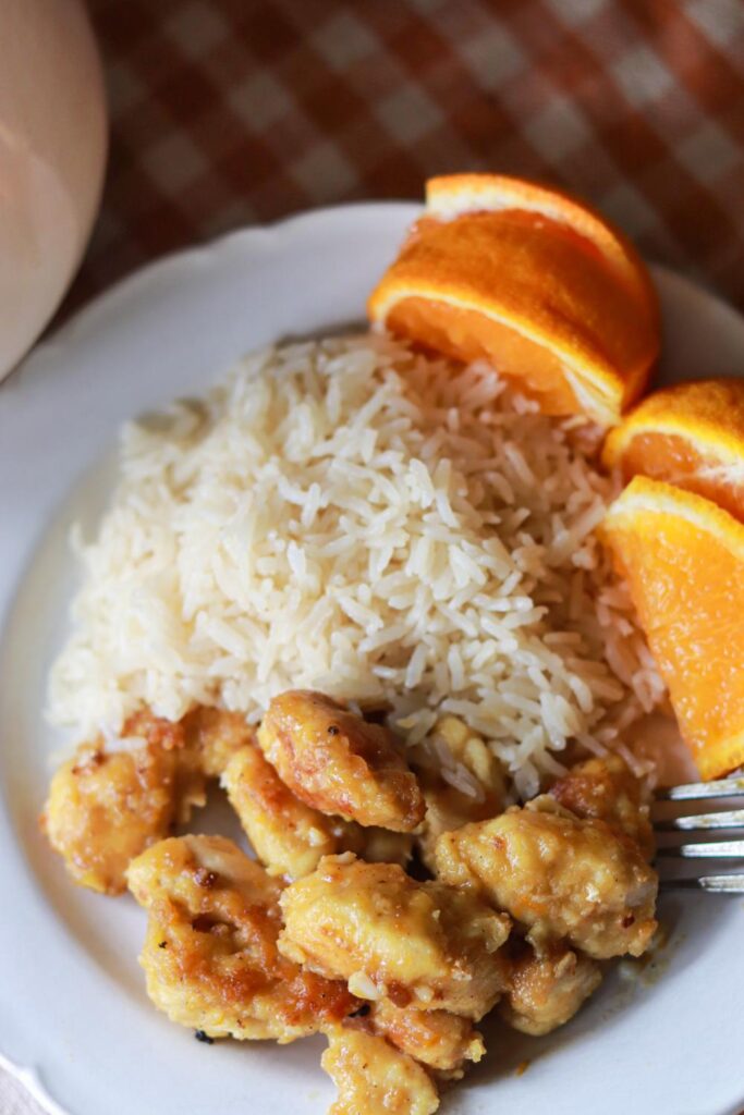 Orange chicken on a white plate with rice and orange slices and a fork.