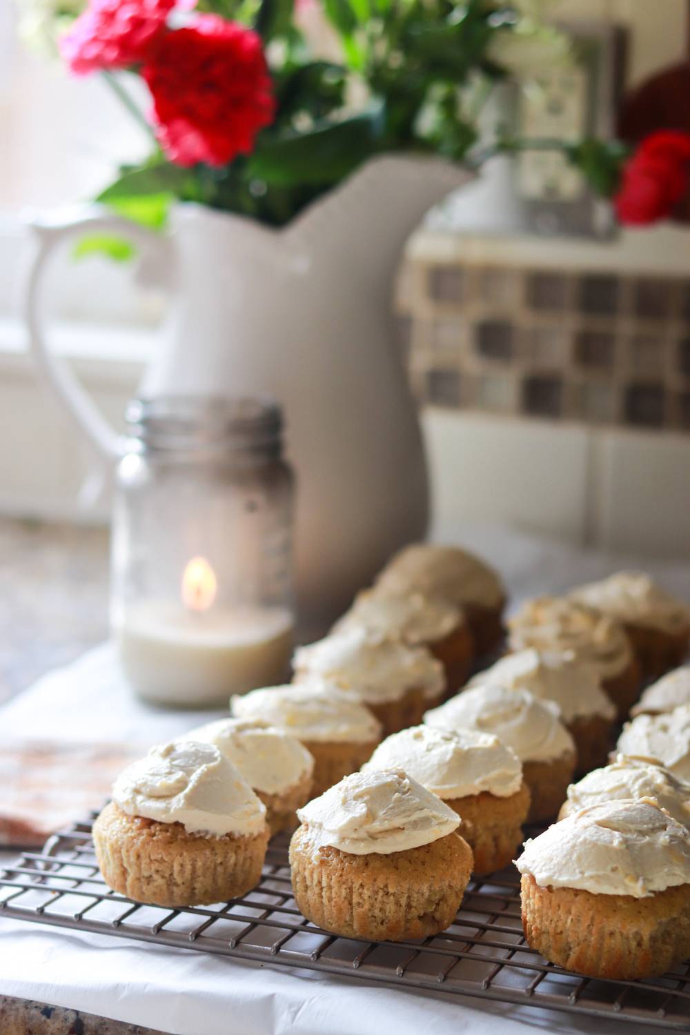 Frosted vanilla cupcakes on a cooling rack with fresh flowers.