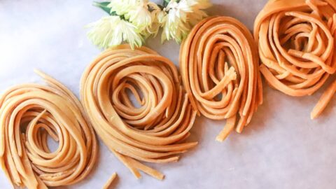Four rolls of homemade pasta lined up on a table with fresh flowers.