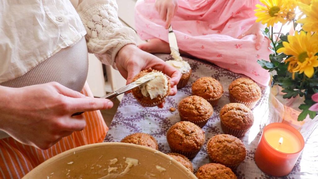 a woman putting cream cheese icing on a cupcake with other plain cupcakes nearby.