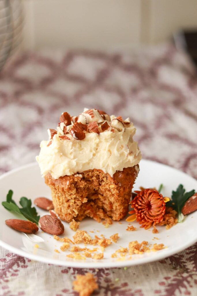 a carrot cake cupcake with cream cheese icing and topped with chopped nuts sitting on a small plate with a bite taken out of it.