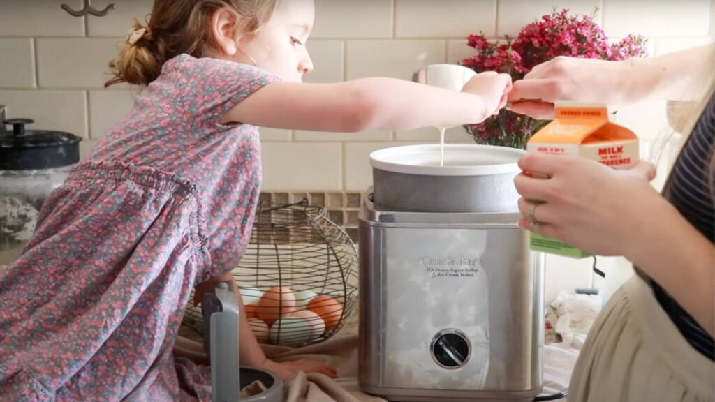 Child helping pour milk into an ice cream maker in a cozy kitchen.
