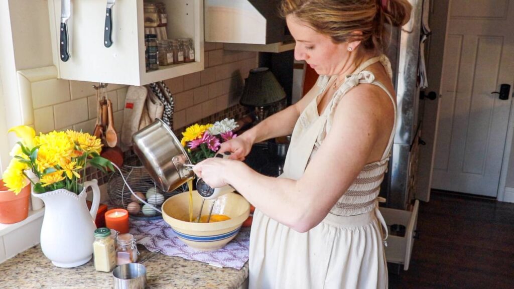 a woman adding melted butter from a small saucepan into a mixing bowl with cupcake batter while using a handheld mixer to combine the ingredients.
