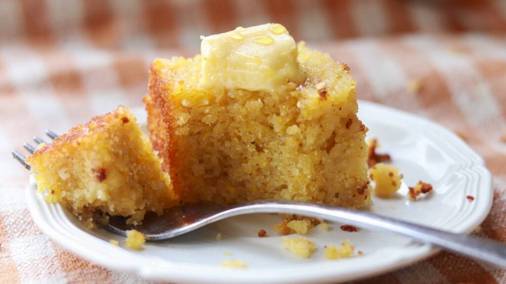 A piece of cornbread with butter on a white plate with a fork.