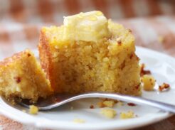 A piece of cornbread with butter on a white plate with a fork.
