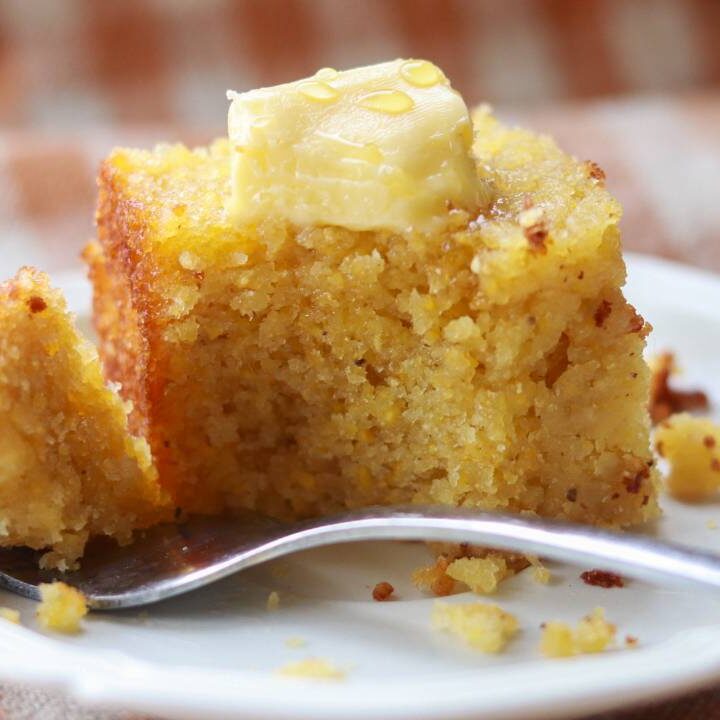 A piece of cornbread with butter on a white plate with a fork.