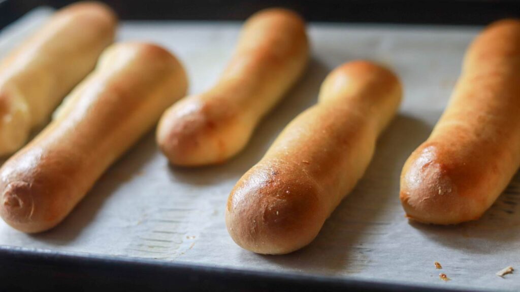 Freshly baked hot dog buns on a baking sheet.