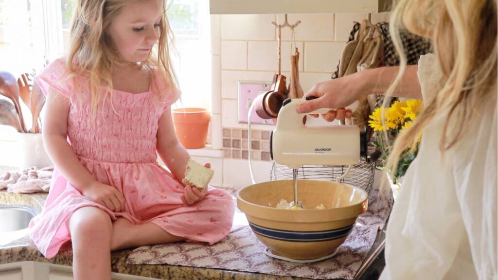 a little girl in a pink dress sitting on a kitchen counter helping her mom make cupcakes using a handheld mixer.